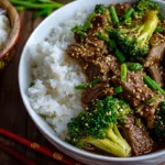 A close-up photograph of a ceramic bowl filled with a finished Chinese beef and broccoli recipe, featuring tender sliced beef and green broccoli florets coated in a glossy brown sauce, with chopsticks resting on the side.