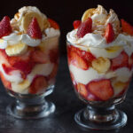 A stunning glass trifle bowl of layered Strawberry Banana Pudding Dream, topped with elegant whipped cream swirls, fresh strawberries, and buttery Chessman cookies.